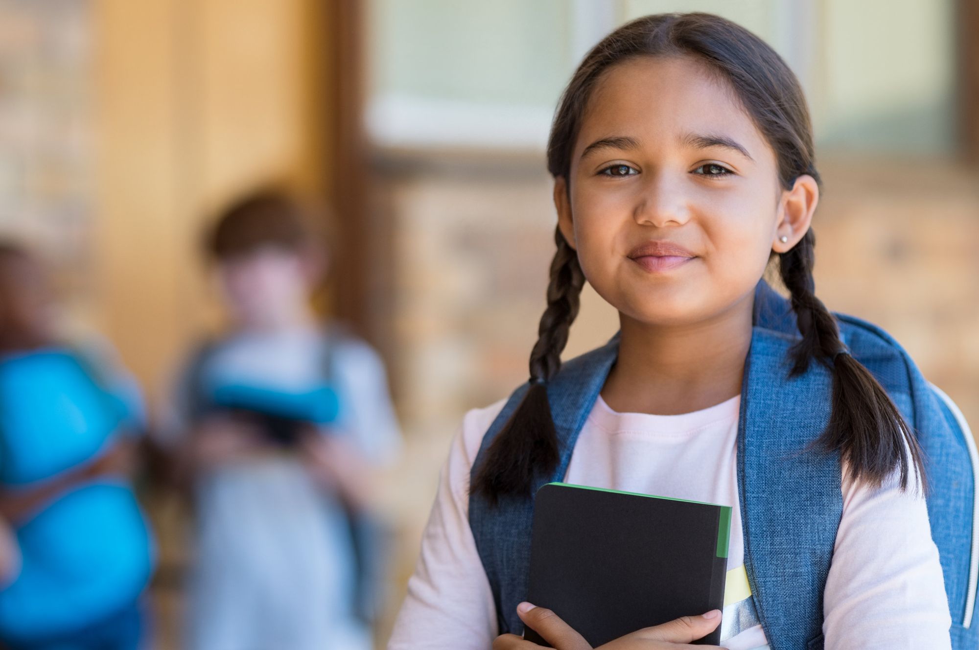 A girl attends school, she has her hair in plaits and is wearing a backpack and carrying a notebook.