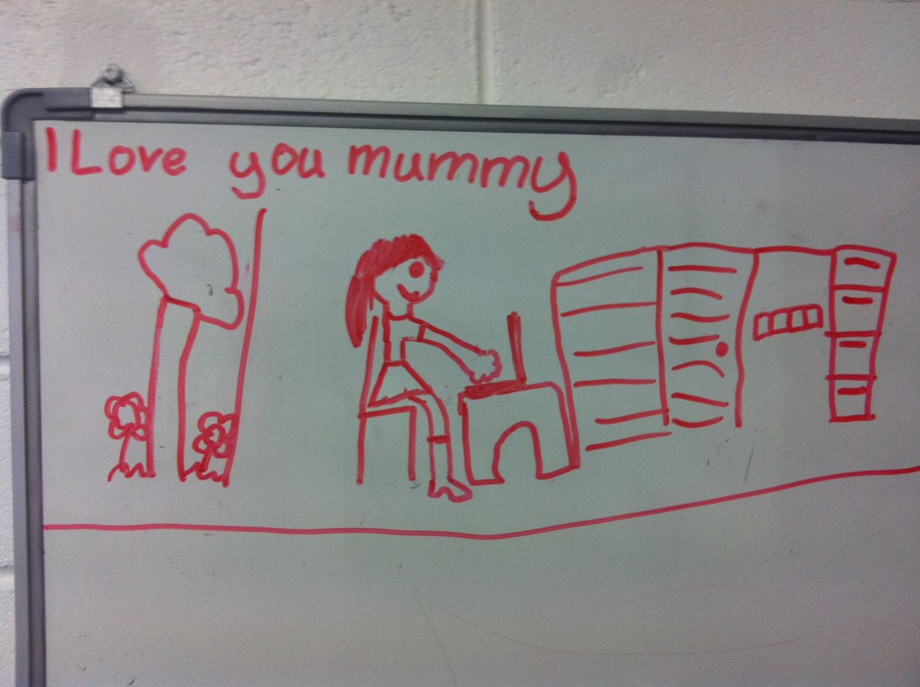 A child's drawing of a woman working at a desk. There's a tree and some flowers. Handwritten text says 'I love you mummy'