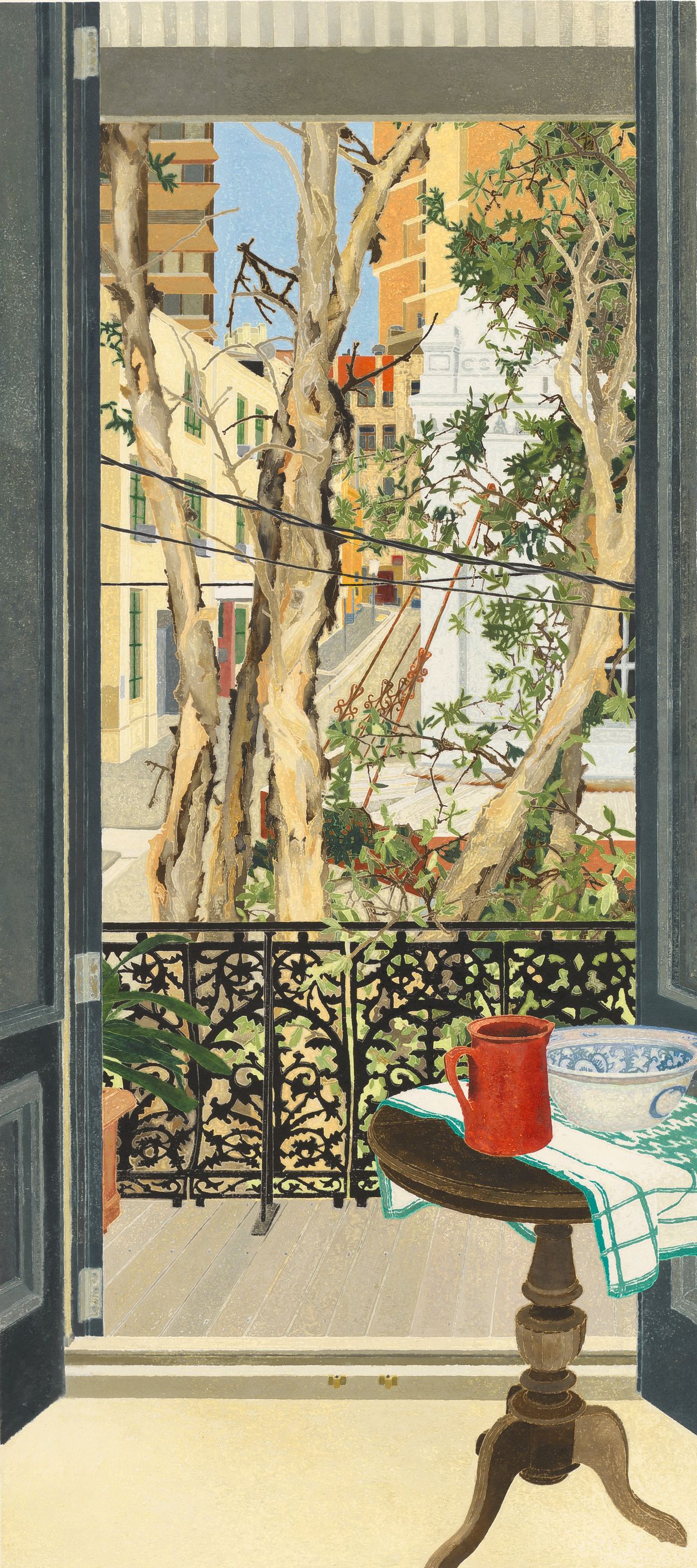 A door opens out onto a balcony. The view takes in trees and an inner urban street. A red jug sits on a table.