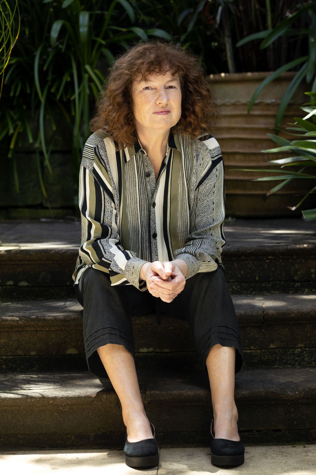 Cressida Campbell sits on some steps in her garden. She is wearing a striped shirt, black trousers and black shoes. She has curly red hair.