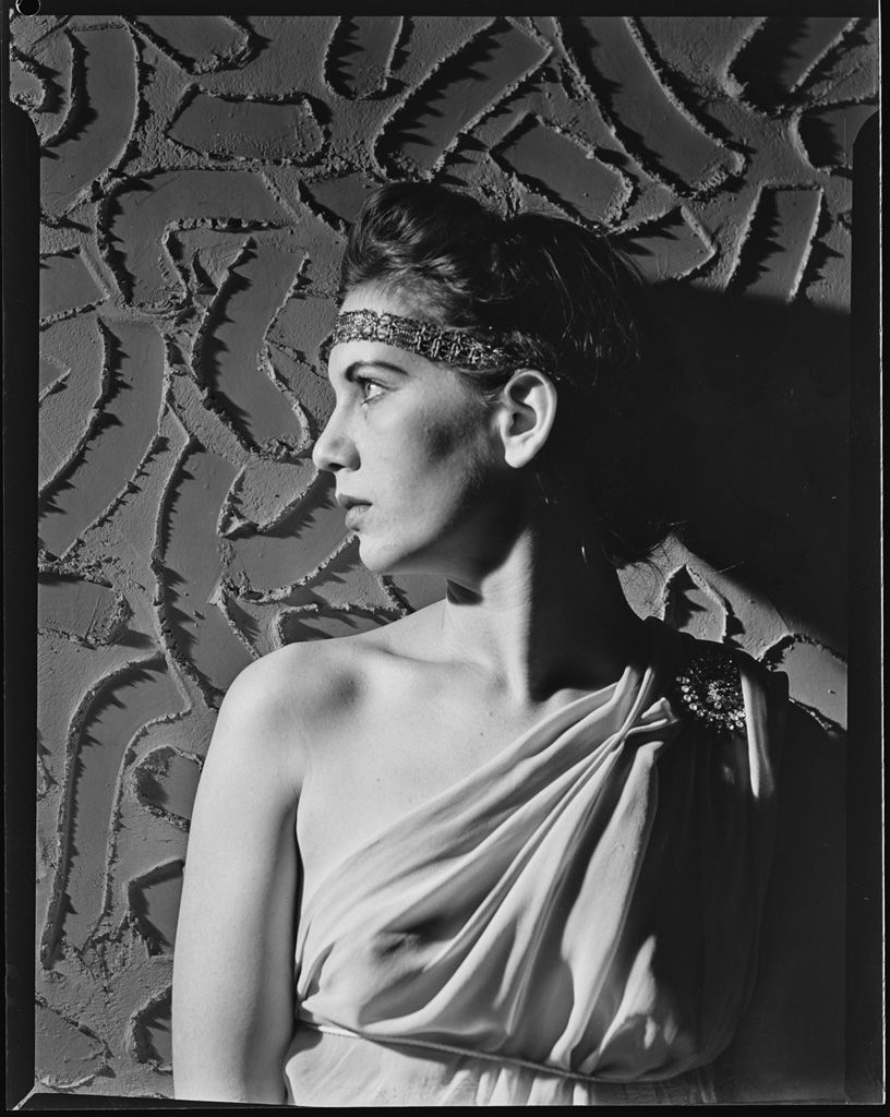 A black and white image of Charmian Clift in a toga-style outfit, against a textured wall. This was a Studio Portrait in 1941 taken by Frederick Stanley Grimes