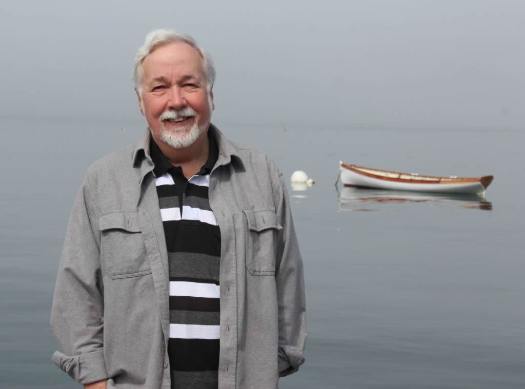 Artist Duncan Ball in front of a grey ocean, with a small fishing boat moored behind him.