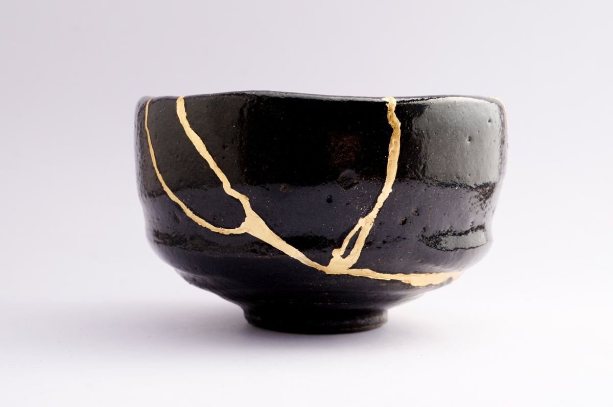 A black Japanese bowl with an uneven shape. It has large cracks in it, fixed with liquid gold.