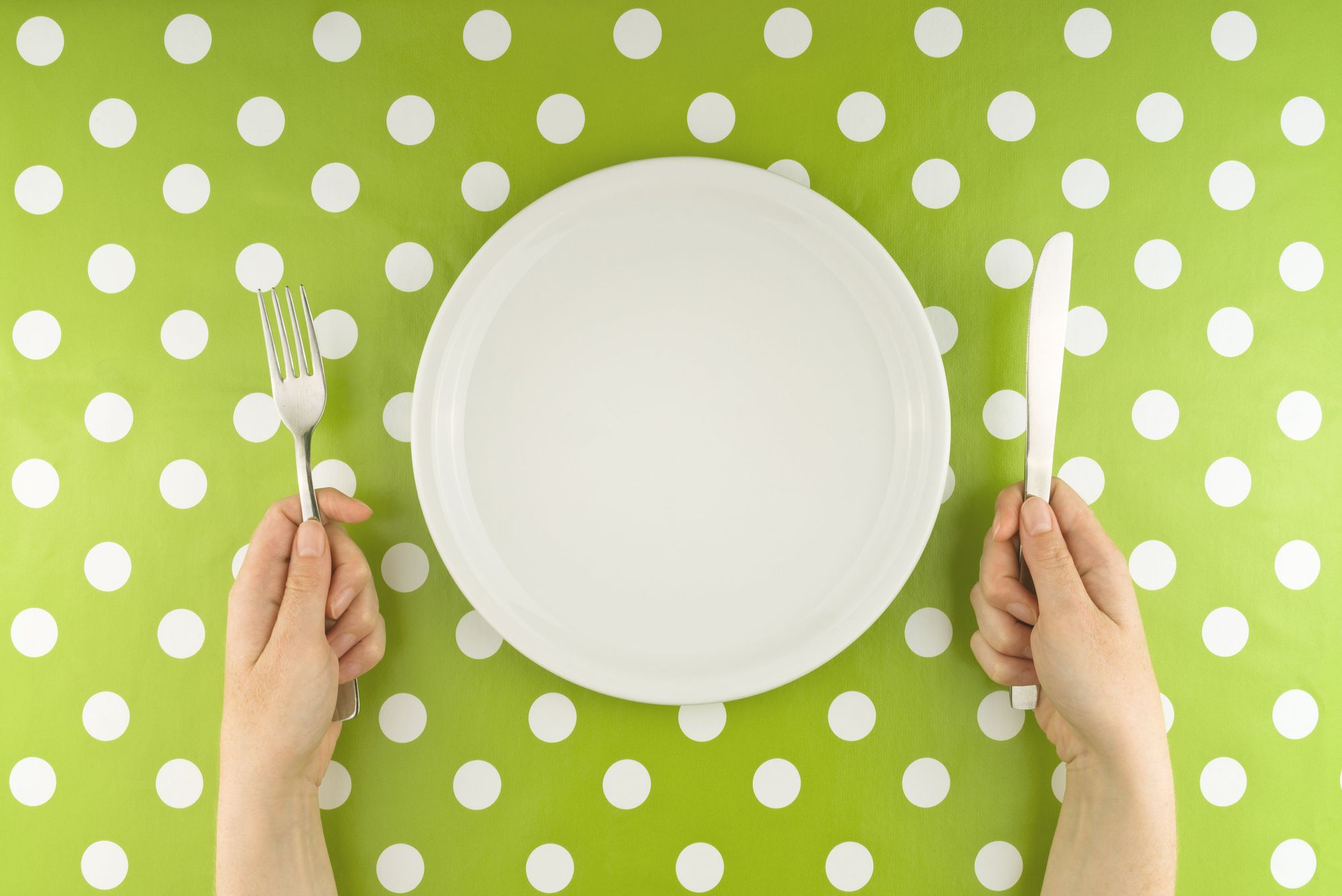 A tablecloth with a green background and white spots. Hands holding cultlery next to an empty white plate.