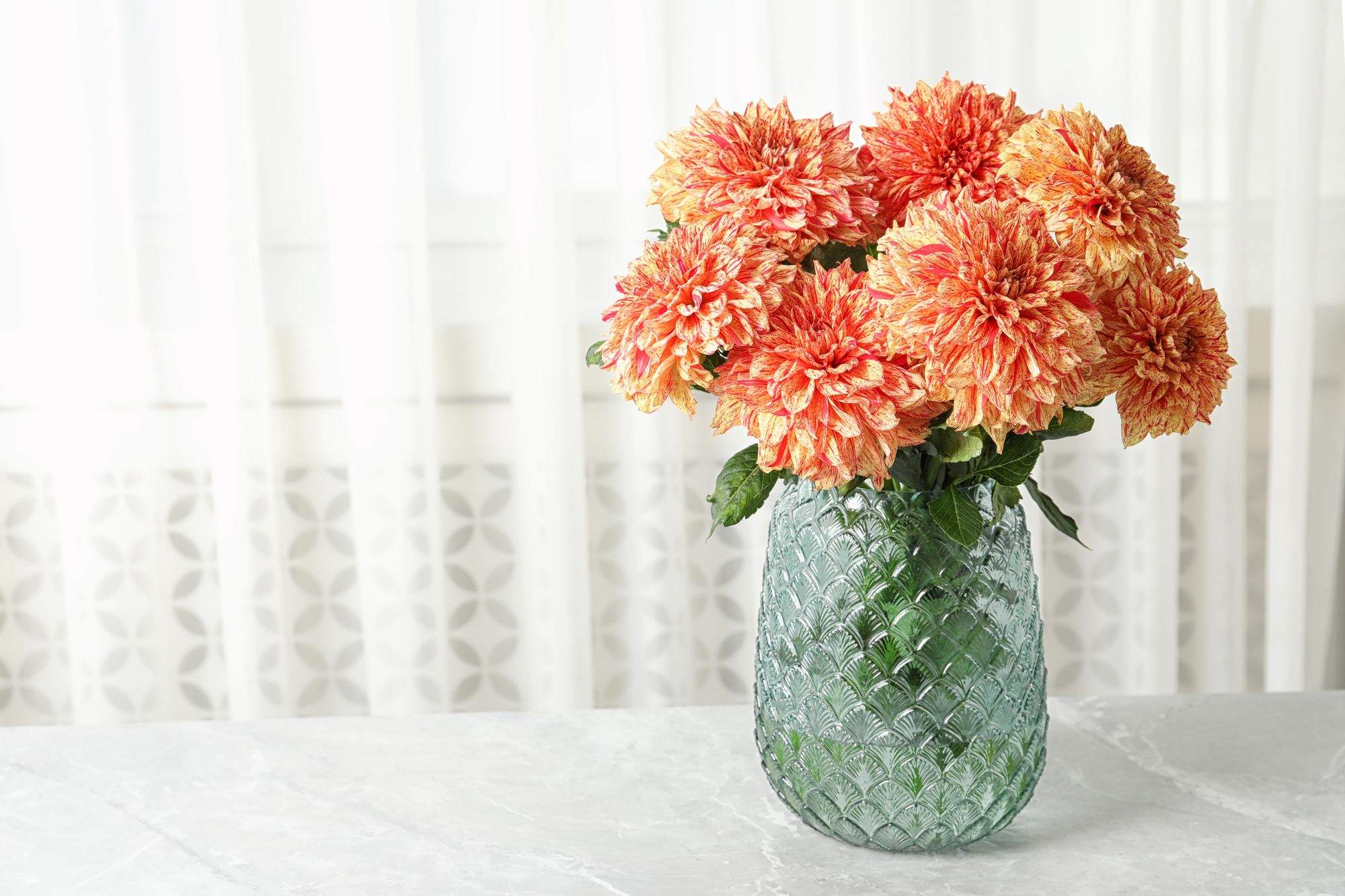 A bunch of orange dahlias in a glass jar on a white table. There's a white curtain in the background. 