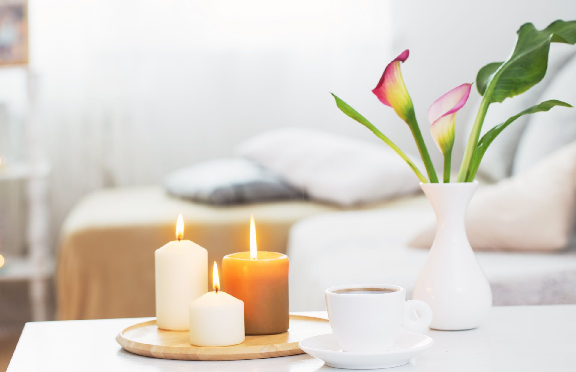 A table with candles, a cup of coffee and a vase of pink arum lillies. There is a couch in the background. 