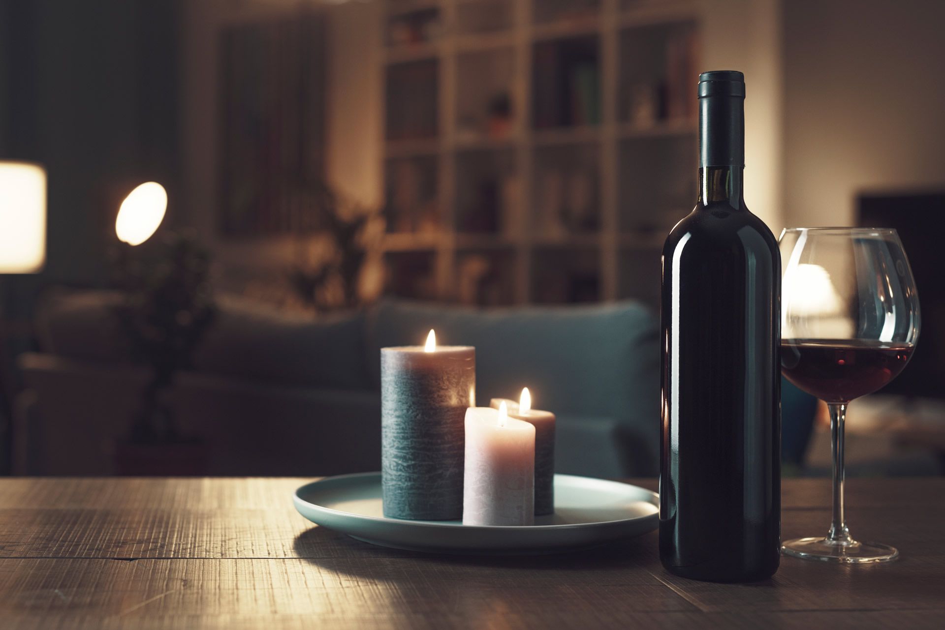 A bottle of wine on a table with some candles in a loungeroom during the evening. 