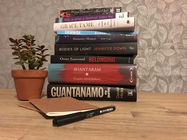 A pile of books on a table with a plant, some notebooks and a pen. 