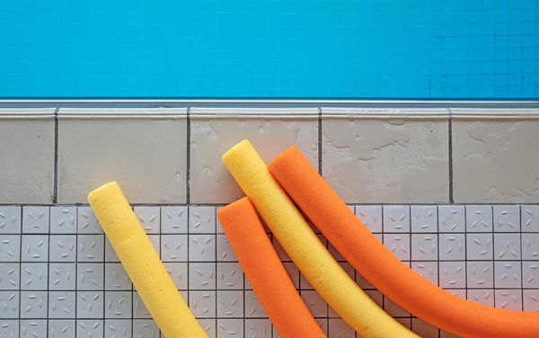 The tiled edge of a swimming pool with four yellow and orange swimming noodles laying on it. 