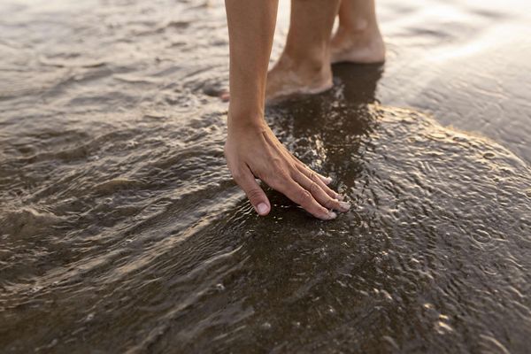 A person leaning over to touch the water at the beach, showing a hand and feet. 